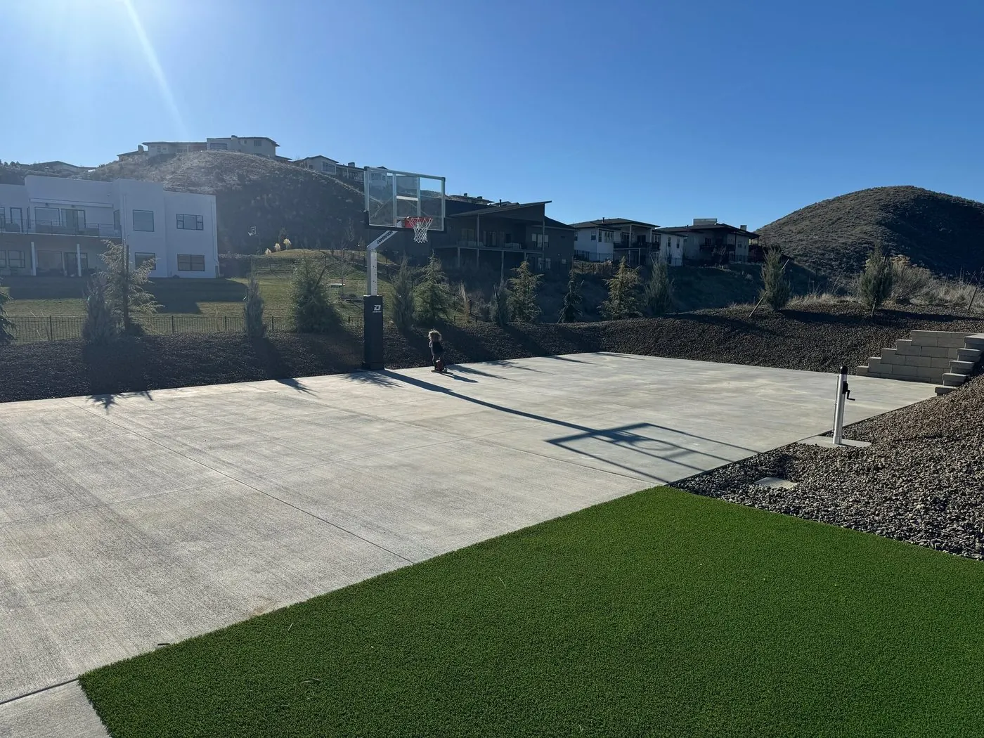 A concrete basketball court with a glass backboard, set into a graded foothills lot between rock-mulched slopes and young evergreens, with turf in the foreground and homes on the hills beyond
