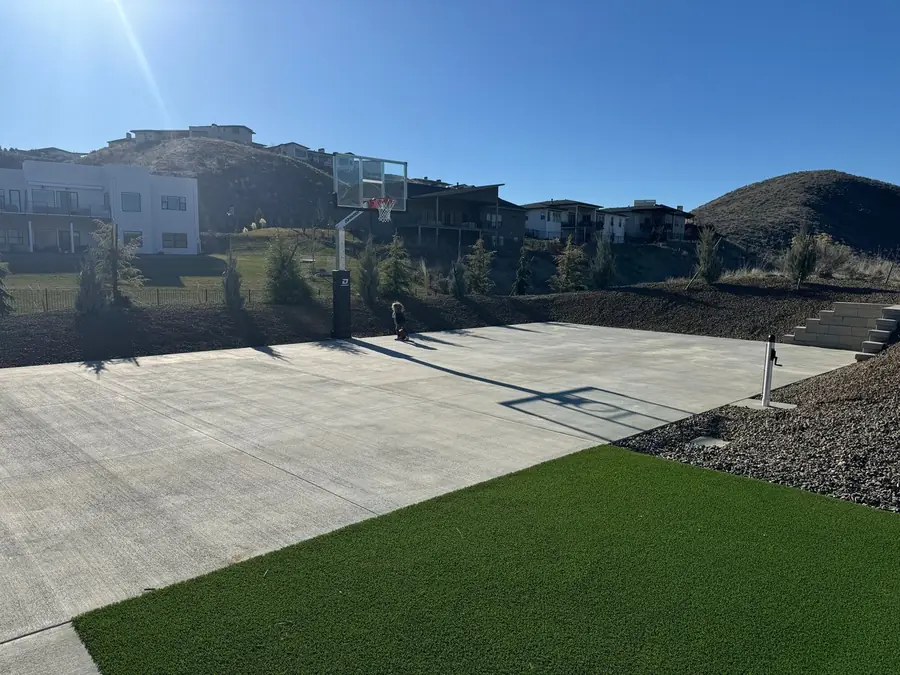A concrete basketball court with a glass backboard, set into a graded foothills lot beside new turf