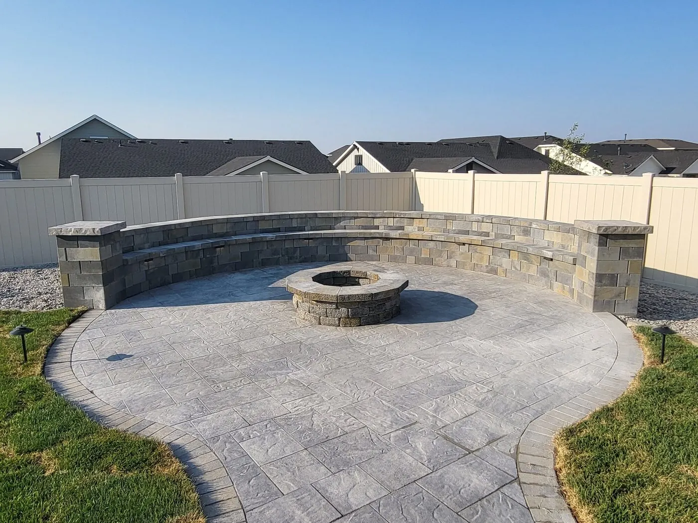 A round stamped-concrete court with a stone fire pit at its center and a curved block seat wall with pillars, in a fenced backyard among new homes
