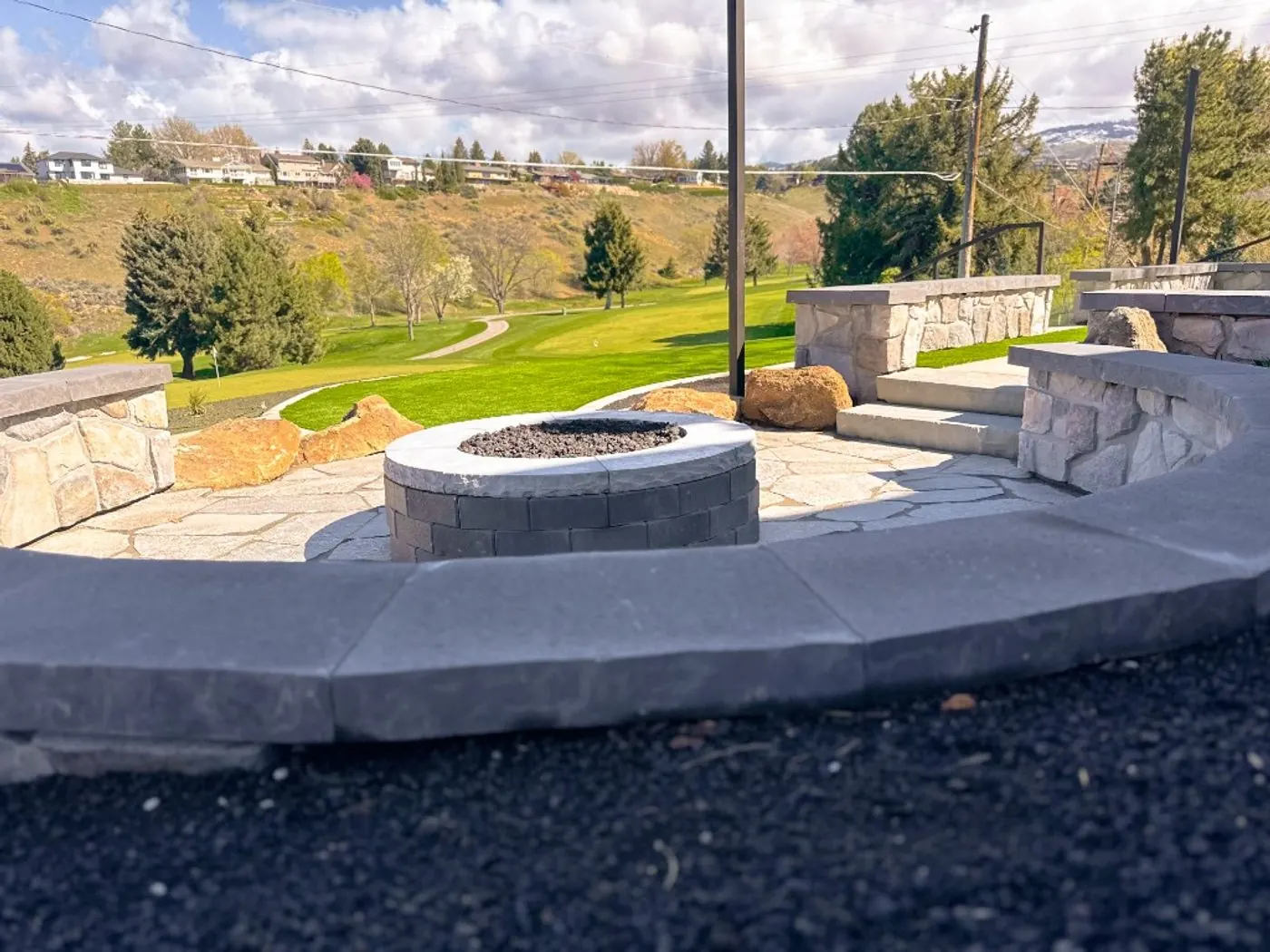 A round gas fire pit with a granite cap on a flagstone patio, wrapped by a curved stone seat wall and looking out over the Crane Creek fairways to the foothills