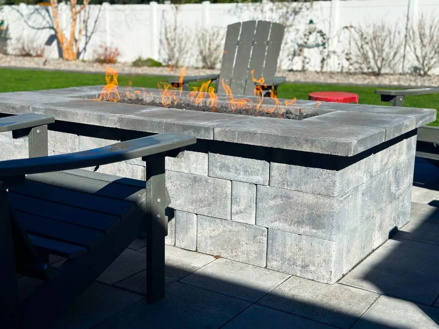 A rectangular gas fire table of stacked gray block under wide concrete caps, with flames over black lava rock and Adirondack chairs pulled up to it