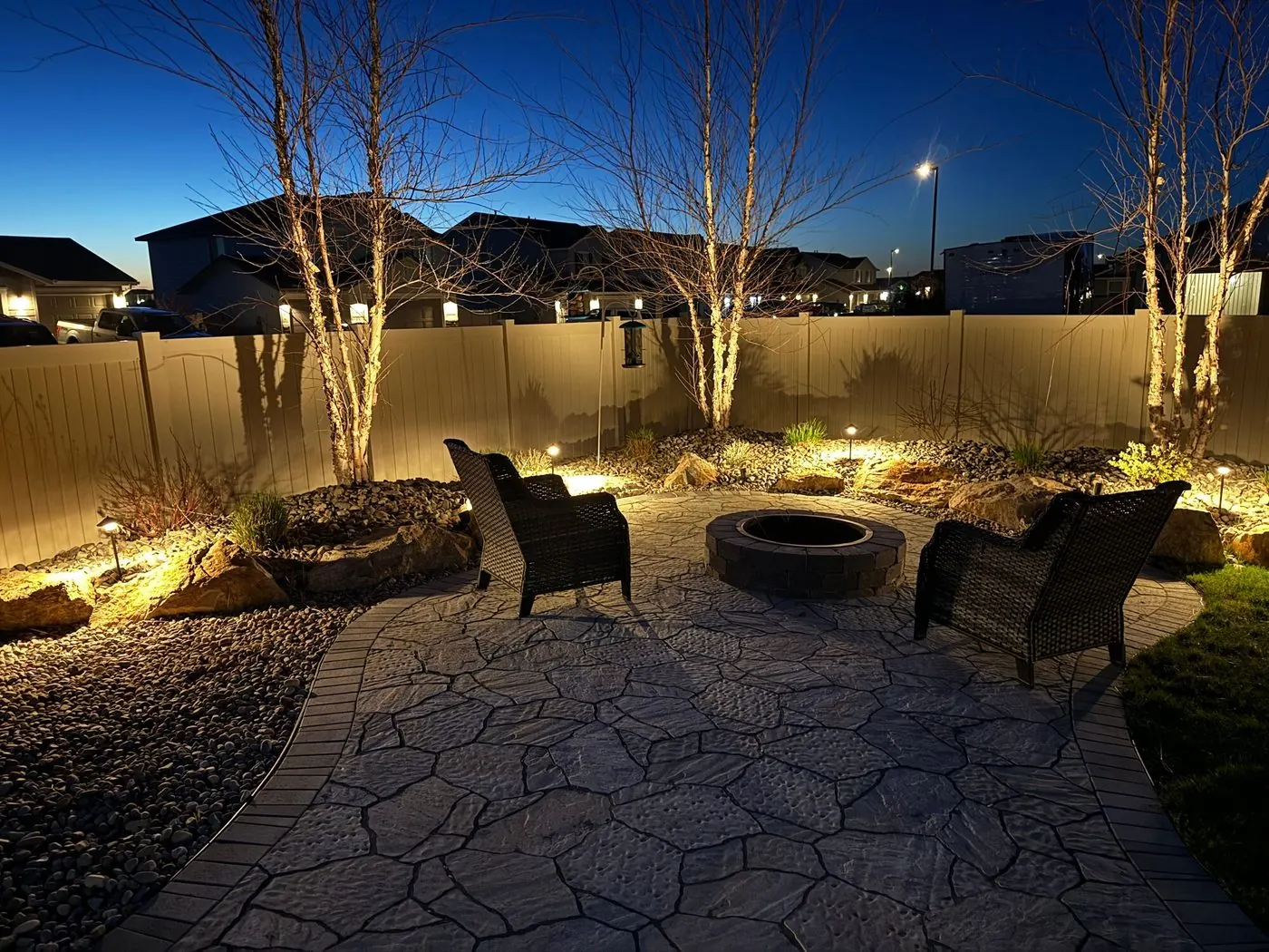 A flagstone-pattern paver terrace at dusk, with two wicker chairs at a round block fire pit and uplit birches along river-rock beds