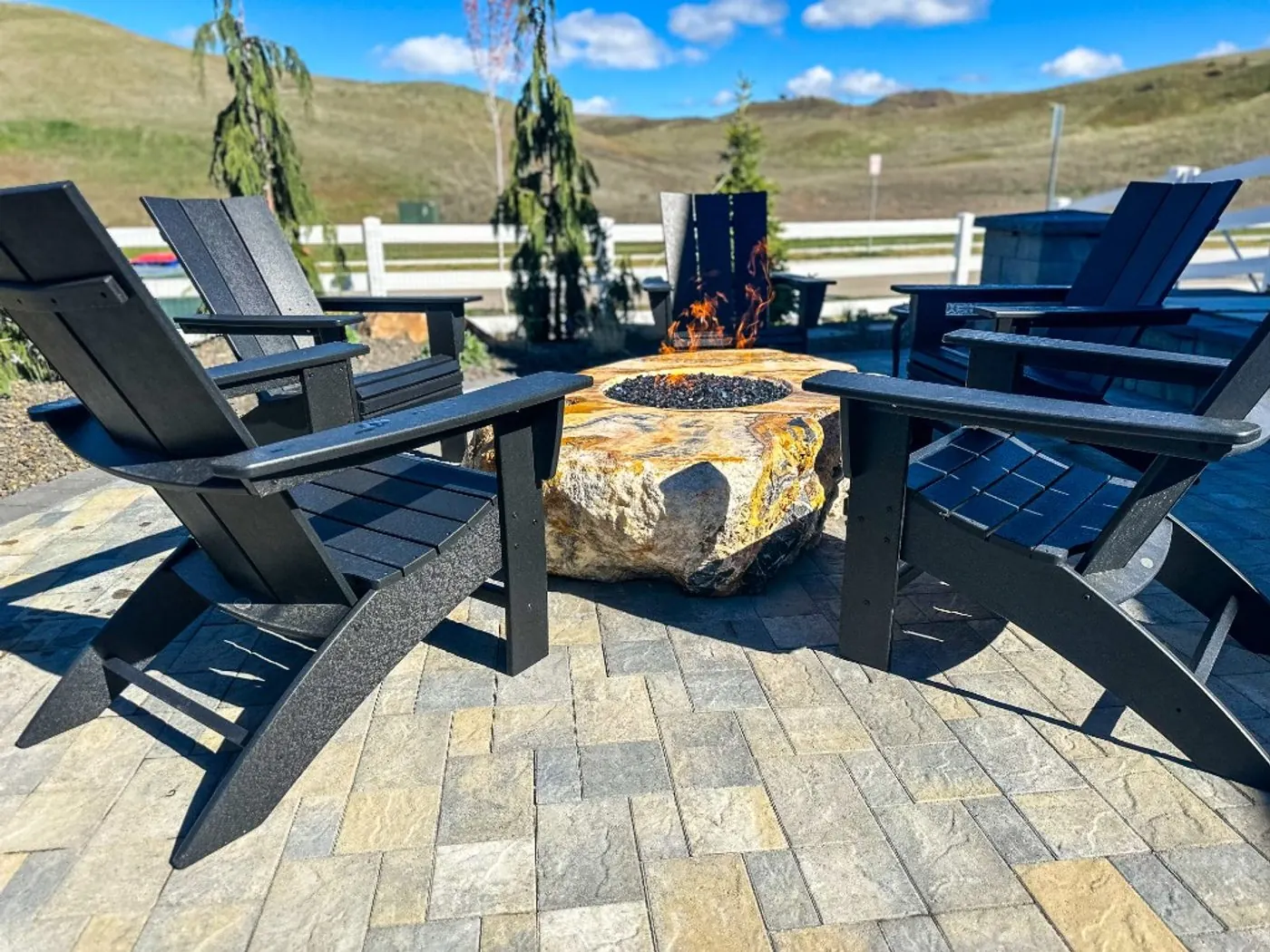Black Adirondack chairs around an amber onyx boulder gas fire pit on slate-blend pavers, with a white rail fence and the Eagle foothills beyond