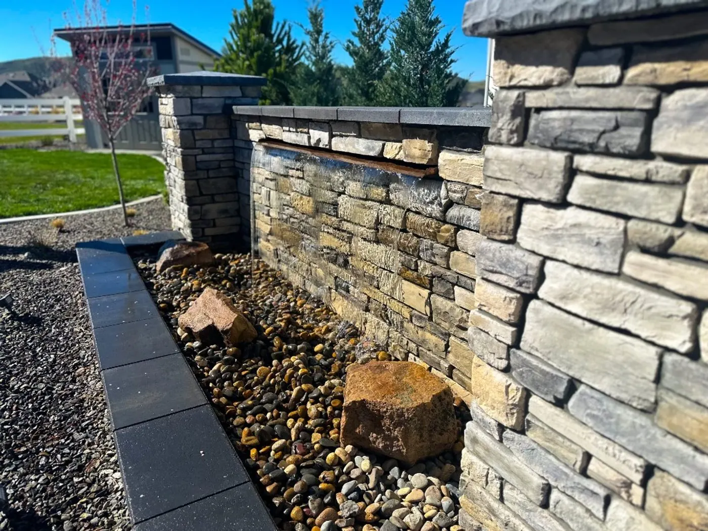 A stacked ledger-stone waterfall wall between slate-capped pillars, its blade spillway sheeting water into a river rock basin edged in black pavers