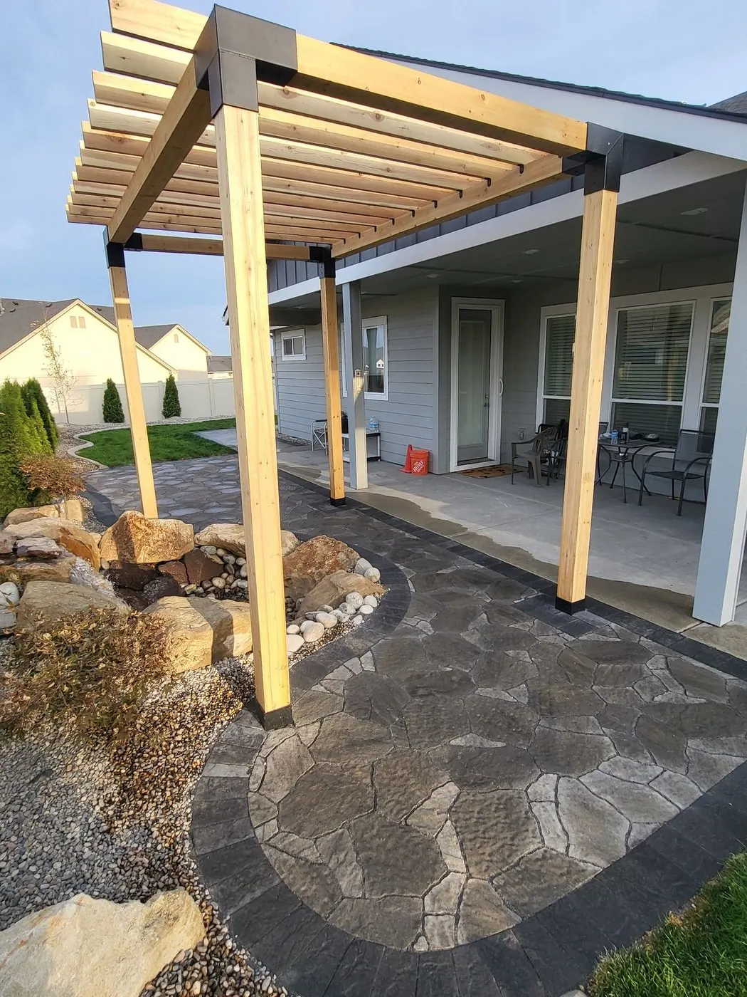 A wood pergola with black brackets over a flagstone-pattern paver terrace beside a covered patio